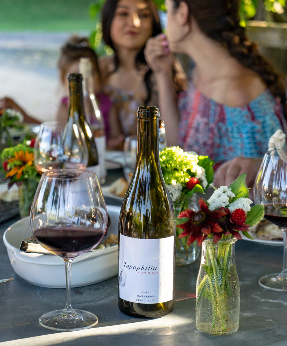Summer dinner party scene featuring Topophilia Wines' Pinot Noir, with a label designed by Limantour Design Studio, elegantly displayed on a festive table setting, embodying the perfect blend of sophistication and warmth for a memorable summer gathering.